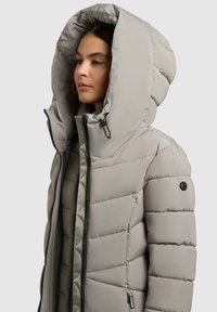 Hellgraue Pufferjacke mit großer Kapuze, gestepptem Design, Frontreißverschluss und einem Logo am linken Arm. Innenfutter in einem dunkleren Farbton.