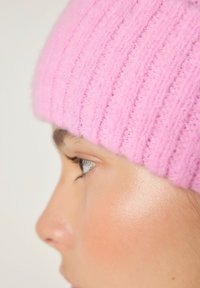Rožinė megzta kepurė su dantytyrinio rašto faktūra, minkšta medžiaga ir prisitaikantis dizainas. Kepurės artimas planas kartu su žmogaus veido šoniniu profiliu.
