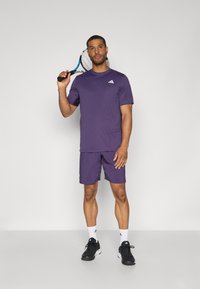 Lila idrottströja och shorts i lättviktsmaterial, med en logotyp på bröstet. Modellen håller en tennisracket.