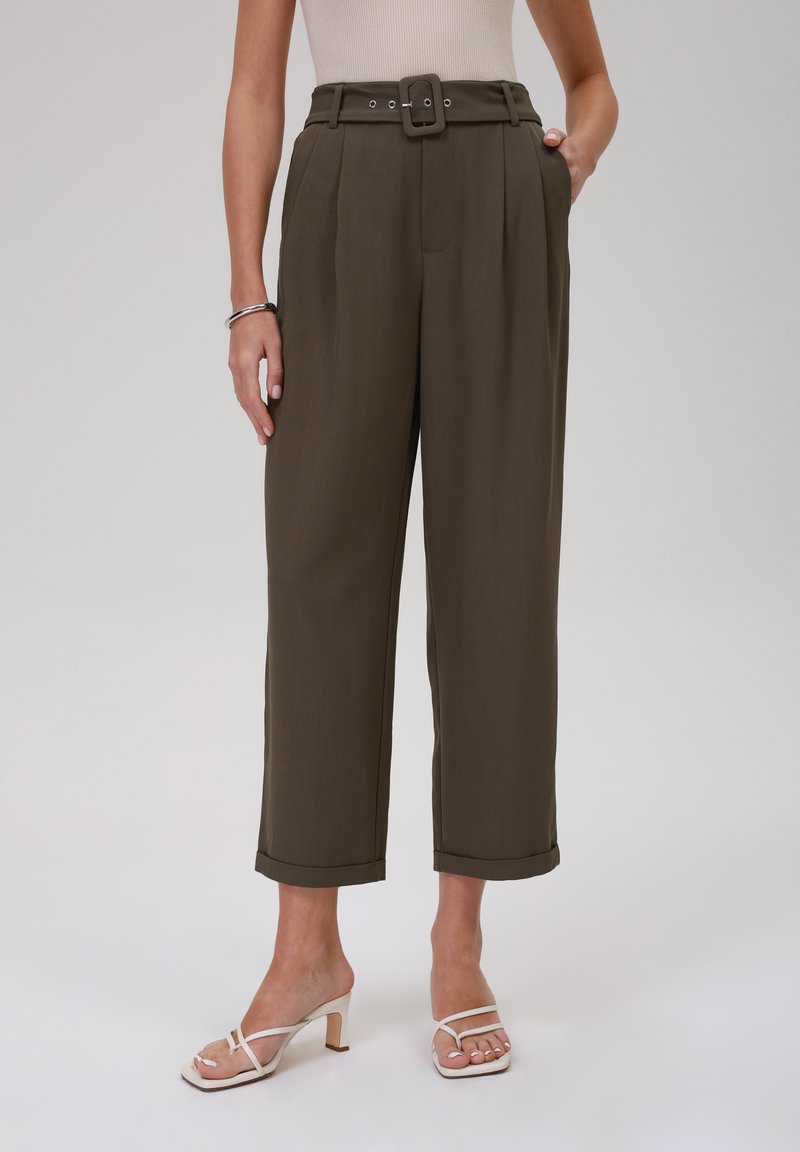 Pantalons taille haute, vert olive, avec une ceinture, des plis et des ourlets retournés ; associés à des talons ouverts à lanières blancs.