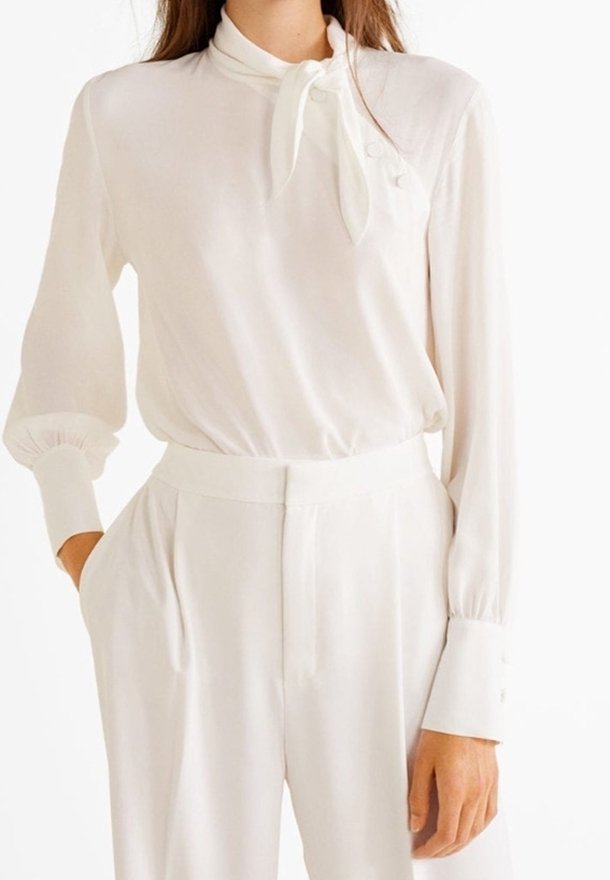 Blouse blanche à manches longues avec un nœud cou et des accents de boutons, associée à un pantalon blanc taille haute. Tissu lisse et léger.