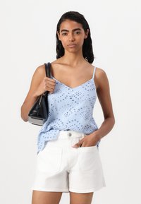 Top sem mangas de cor azul claro com padrões de ilhós, decote em V e um corte descontraído; combinado com calções brancos e uma mala preta.