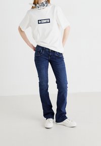 T-shirt blanche avec logo Converse marine, jean en denim bleu et baskets blanches. Modèle portant un foulard à motifs noir et blanc autour du cou.