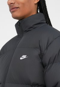 Fekete puffermellény magas gallérral, cipzáras záródással és fehér Nike logóval a mellkason. Simára csiszolt textúra, steppelt kialakítás.