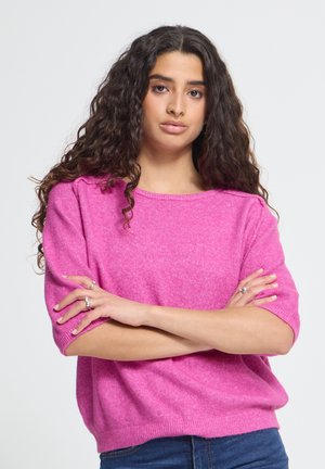 Vrouw met lang krullend haar die een felroze trui en blauwe spijkerbroek draagt, staat met gekruiste armen tegen een effen achtergrond.