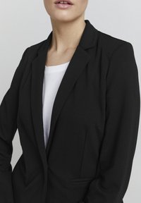 Černý blazer se fitted designem, klopami a p ředními kapsami. Nosič má na sobě bílý tričko, které se vyznačuje hladkou texturou a upraveným siluetou.