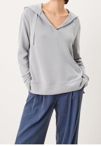 Sudadera con capucha de color gris claro, con un diseño de cuello en V y mangas largas. Cuenta con cordones ajustables y un corte relajado. Combinada con pantalones azules.