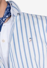 Camisa de botones de rayas en blanco y azul claro con un collar blanco y un pequeño diseño bordado en el lado izquierdo del pecho. Tejido texturizado.