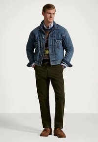 Hombre vistiendo una chaqueta de mezclilla sobre un suéter de estampado y una camisa con cuello, pantalones oscuros y zapatos de ante marrones, de pie con las manos en los bolsillos.