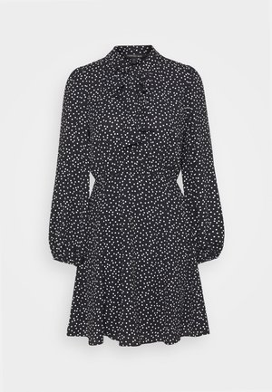 Vestido negro con lunares blancos, mangas largas abullonadas, cintura fruncida y un lazo atado en el cuello.