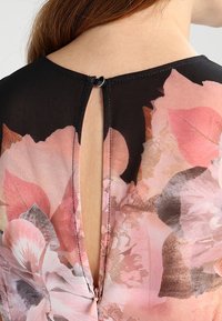 Blusenshirt mit floralem Druck aus transparentem, schwarzem Stoff, mit einer Schlüssel-Ausschnitt-Rückenschließe und zarten rosa und beige Blumenmustern.