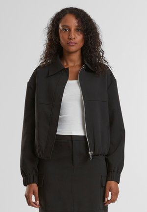 Vrouw met krullend haar draagt een zwarte jas met rits over een wit geribd tanktopje en een zwarte rok met voorzakken, staand tegen een effen achtergrond.