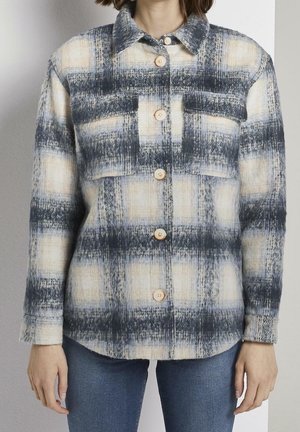 Frau trägt ein beige-grau kariertes Hemd mit Knöpfen, zwei Brusttaschen und blaue Jeans, steht vor einem einfarbigen Hintergrund.