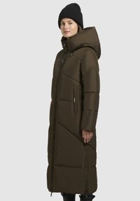 Žena oblečená v dlhej tmavohnedej zimnej prešívanej bunde s kapucňou a čiernou pletenou čiapkou, stojí proti jednouchému svetlošedému pozadiu.