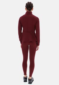 Vínová peplum bunda s vysokým límcem spojená s přiléhavými vínovými legínami. Hladká textura a na míru šitý design s jemnými švy.