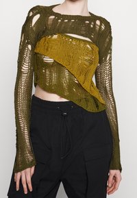 Strikket cropped sweater i jordlige oliven- og sennepsfarver, har et lagdelt design med udskæringer og teksturerede mønstre. Sammen med sorte cargo bukser.