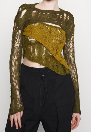 Strikket cropped sweater i jordlige oliven- og sennepsfarver, har et lagdelt design med udskæringer og teksturerede mønstre. Sammen med sorte cargo bukser.