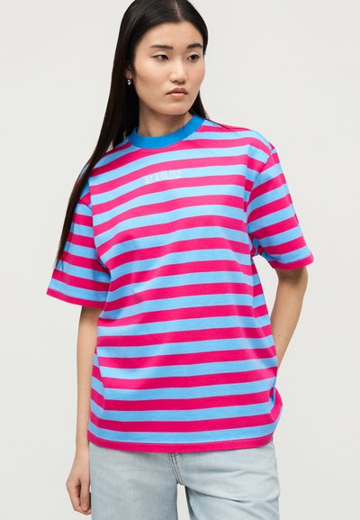 Γυναίκα που φορά μπλε και ροζ οριζόντια ριγέ κοντομάνικη μπλούζα με το κείμενο «STIEGLITZ», συνδυασμένη με ανοιχτόχρωμο τζιν, ποζάρει σε απλό φόντο.