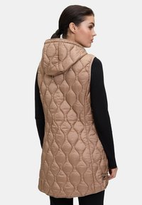 Gilet imbottito beige con cappuccio, caratterizzato da un motivo trapuntato e un taglio lungo. Texture liscia e dettaglio hardware sui lati. Maglietta nera a maniche lunghe.