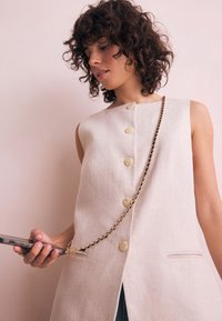 Bēšs lina vestes ar piecām lielām pogām, sānu kabatām un pievienotu zelta ķēdi telefona turēšanai. Gluda tekstūra, minimālistisks dizains.