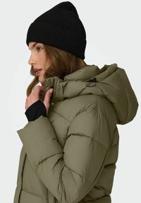 Olivgrüne, isolierte Jacke mit fluffigem Design und einer Kapuze, kombiniert mit einer schwarzen Strickmütze. Nahaufnahme, die Textur und Passform hervorhebt.