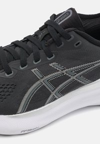 Zwart sportief schoen met geoefend bovenwerk van stof, reflecterende zilveren accenten en gevoerde witte zool. Voorzien van veters en merkopdruk op de tong.