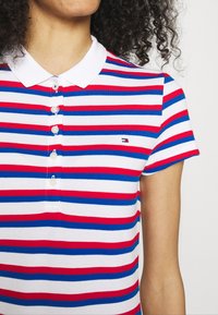 Randig polo skjorta i vitt, rött och blått. Har krage, fem knappar och en liten logotyp. Mjuk, strukturerad tyg. Korta ärmar.