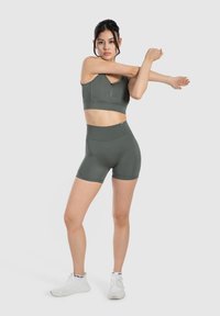 Sujetador deportivo verde y pantalones cortos de cintura alta en tejido elástico. Presenta un detalle con cremallera en el sujetador. Modelo lleva zapatillas deportivas blancas.