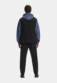 Chaleco acolchado negro sobre una sudadera azul, combinado con pantalones negros. Textura suave, acabado ligeramente brillante, diseño minimalista, características ajustables.