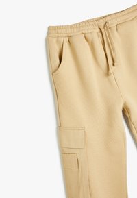 Beige cargo bukser har en elastisk talje med snor, sidelommer og en lappetaske på ydersiden af låret. Fremstillet af blødt stof.