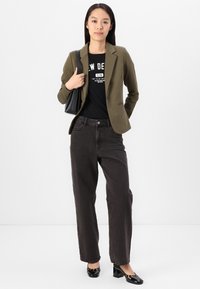 Kvinna stående i olivgrön kavaj, svart grafisk t-shirt, lösa svarta jeans, glansiga svarta klackar, hållande svart axelväska mot vit bakgrund.