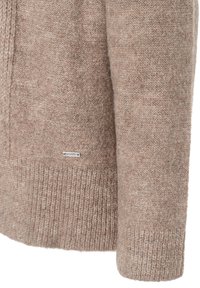 Beige gestrickter Pulloverärmel und -bund mit geripptem Saum und kleinem rechteckigen Markenschild nahe der unteren Kante.