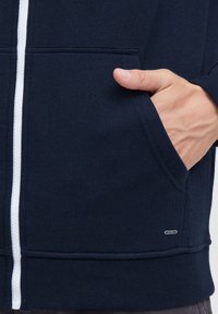 Sudadera azul marino con cremallera hecha de un tejido suave, con un bolsillo canguro, que presenta una cremallera blanca y una pequeña etiqueta de la marca en el dobladillo.