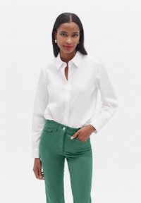 Camicia bianca con bottoni e maniche lunghe, abbinata a pantaloni verdi aderenti. La camicia presenta un colletto a punta e una leggera lucentezza.