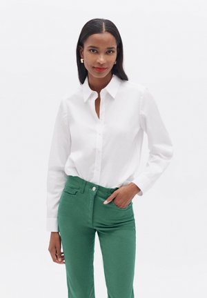 Chemise blanche à manches longues, associée à un pantalon vert ajusté. La chemise présente un col pointu et une légère brillance.