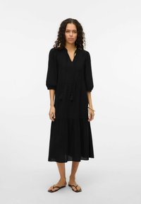 Svart knälång klänning i lätt tyg. Har puffärmar med 3/4 längd, en V-ringning och dragsko med tofsar. Utställd nederkant.