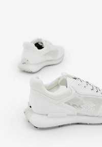 Zapatillas blancas con cordones que presentan una parte superior de cuero suave con acentos de malla, una suela acolchada y un logotipo texturizado en el lateral.