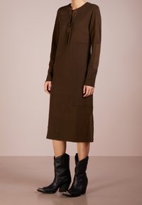 Abito lavorato a maglia marrone con maniche lunghe, spalle scese e chiusura frontale con bottoni. Abbinato a stivali cowboy neri alla caviglia con un accento argentato.