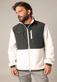 Homme avec une barbe portant une veste zippée bicolore en blanc et vert foncé, se tenant les mains relaxées, regardant sur le côté devant un fond uni.