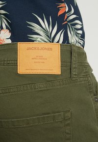 Pantalones verdes con etiqueta de cuero que dice "JACK&JONES," con detalles de costura y bolsillos. El fondo muestra una camisa de patrón floral oscuro.
