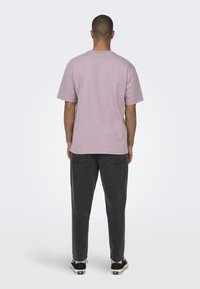 Camiseta de manga corta morada claro, material de algodón, corte relajado, combinada con pantalones gris oscuro, diseño minimalista, sin logotipos visibles.