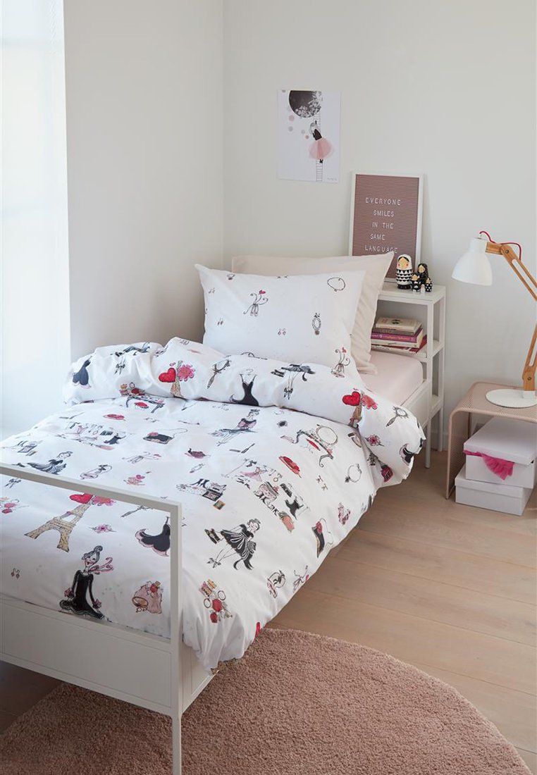 Ein einzelnes weißes Bett mit einem Paris-Themen-Bettbezug, ein kleiner Nachttisch mit einer Lampe und Büchern, zwei Puppen, Wandkunst und einem runden rosa Teppich.