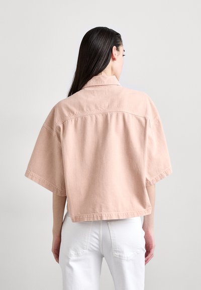 Rožinė džinsinė striukė trumpomis rankovėmis, dėvima laisvai, su apykakle. Yra šiek tiek nuplauta tekstūra ir matomi siuvimo detalės. Po ja dėvimi balti džinsai.