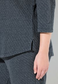 Mano di una persona appoggiata accanto all'anca, indossando pantaloni blu scuro con trama e un top abbinato con una piccola apertura laterale.