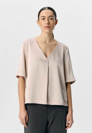 Blusa a maniche corte con scollo a V in tessuto beige chiaro, caratterizzata da una silhouette morbida e un delicato dettaglio plissettato sul davanti, abbinata a pantaloni scuri.