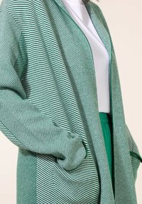 Cardigan rayé vert et blanc à coupe ample, manches longues et deux poches latérales. Fabriqué en tissu doux et léger.