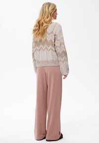 Vrouw met lang blond haar draagt een beige gebreide trui met patroon en losse stofroze broek, staande met de gezicht naar rechts op een plain witte achtergrond.