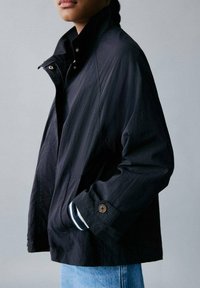 Chaqueta negra ligera con cuello alzado, cierre de cremallera y puños ajustables. Presenta una textura sutil y acentos de rayas en contraste.