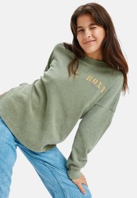 Jonge vrouw in een groene Roxy-trui en blauwe jeans knipoogt terwijl ze naar voren leunt tegen een witte achtergrond.