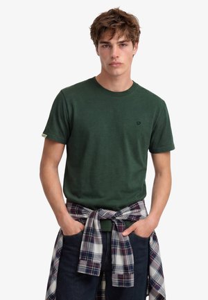 Junger Mann trägt ein dunkelgrünes Kurzarmhemd und ein kariertes Langarmhemd, das um die Taille gebunden ist, Hände in den Jeans-Taschen.